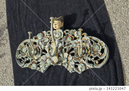 XALO, SPAIN - JUNE 20, 2026.A vintage wall coat hanger shows detailed metal carvings in floral shapes with a hand painted Balinese face at the top. The hanger is on a black surface. XALO, SPAIN - JUNE 20, 2026.A vintage wall coat hanger shows detailed metal carvings in floral shapes with a hand painted Balinese face at the top. The hanger is on a black surface. 141221047