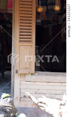 Open wooden shutter reveals shadowed artisan shop interior with hanging lanterns, woven textiles, brick details, painted wood, and warm natural light spilling across worn surfaces Open wooden shutter reveals shadowed artisan shop interior with hanging lanterns, woven textiles, brick details, painted wood, and warm natural light spilling across worn surfaces 141221227