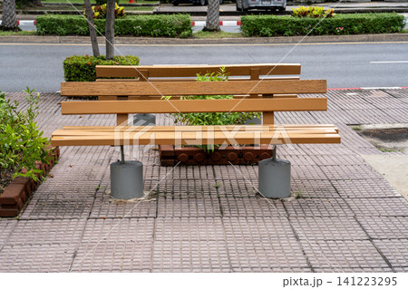Modern wooden bench on a paved sidewalk with concrete bases. Public seating area in an urban park or street setting with green plants and road in the background. Professional outdoor furniture. 141223295