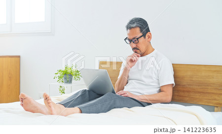 ベッドでノートPCを使うシニア男性。パソコン、仕事、DX、リラックス、ワーケーション、テレワーク ベッドでノートPCを使うシニア男性。パソコン、仕事、DX、リラックス、ワーケーション、テレワーク 141223635