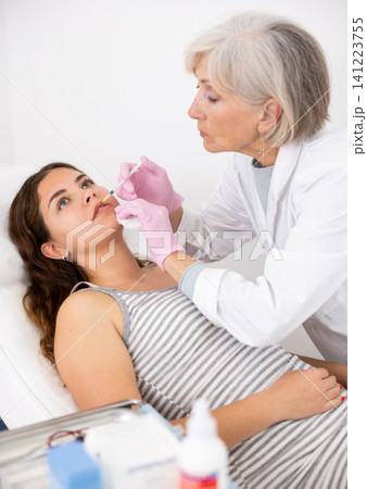 Professional mature female beautician with syringe injecting filler with hyaluronic acid in lips of young woman client during procedure in beauty salon 141223755