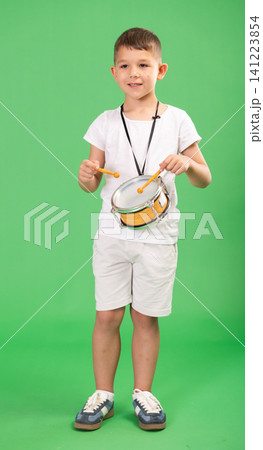 Boy in white t-shirt and shorts drums on children drum that hangs from his neck against green background Boy in white t-shirt and shorts drums on children drum that hangs from his neck against green background 141223854