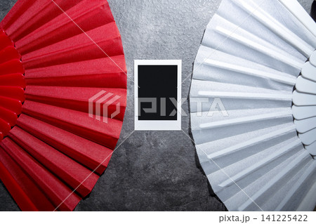 A high-angle studio shot featuring a vibrant red folding hand fan and a white lace fan placed on a grey textured background, with a blank instant film photo frame centered in the middle 141225422