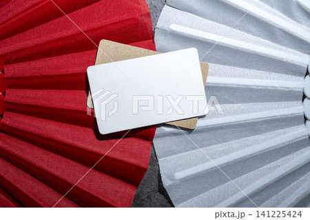A close-up top view of a blank white rectangular card placed on top of a cork surface, framed by textured red and white paper fan pleats, perfect for design mockups A close-up top view of a blank white rectangular card placed on top of a cork surface, framed by textured red and white paper fan pleats, perfect for design mockups 141225424