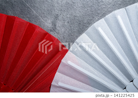 An artistic, high-contrast close-up shot showing the textures and pleated patterns of a red and white folding fan, set against a dark gray textured background An artistic, high-contrast close-up shot showing the textures and pleated patterns of a red and white folding fan, set against a dark gray textured background 141225425