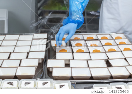 Food factory worker in blue sterile gloves arranging orange gelatin desserts into small square paper containers on a tray in a professional kitchen Food factory worker in blue sterile gloves arranging orange gelatin desserts into small square paper containers on a tray in a professional kitchen 141225466