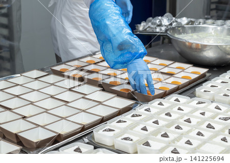 Food factory worker in blue sterile gloves arranging orange gelatin desserts into small square paper containers on a tray in a professional kitchen Food factory worker in blue sterile gloves arranging orange gelatin desserts into small square paper containers on a tray in a professional kitchen 141225694