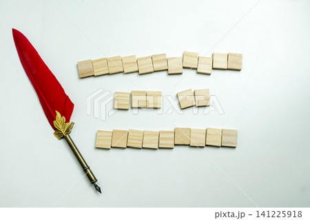A conceptual flat lay featuring a red feather quill pen next to blank wooden tile blocks arranged in lines on a clean white background, symbolizing writing and censorship A conceptual flat lay featuring a red feather quill pen next to blank wooden tile blocks arranged in lines on a clean white background, symbolizing writing and censorship 141225918