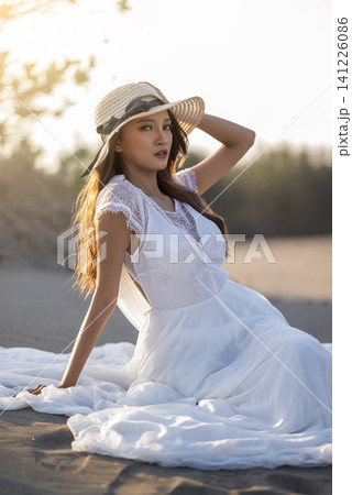 A beautiful young Indonesian southeast asian woman in an elegant white lace dress and straw hat poses gracefully on the sandy beach during golden hour 141226086