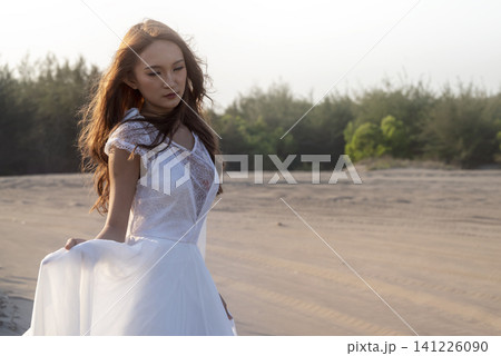 A beautiful young Indonesian southeast asian woman in an elegant white lace wedding dress stands thoughtfully on a sandy beach, looking down with a romantic and peaceful expression A beautiful young Indonesian southeast asian woman in an elegant white lace wedding dress stands thoughtfully on a sandy beach, looking down with a romantic and peaceful expression 141226090
