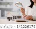 Woman holding magnifying glass, looking at house model on desk while using calculator to analyze finance, search for perfect home loan or plan real estate investment budget 141226179