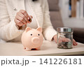 Woman putting coin into piggy bank represents personal finance saving money for future retirement planning with glass jar of coins on table, showing financial growth and responsibility 141226181