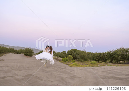 A Indonesian southeast asian romantic couple dressed in white strolling across sandy dunes during a soft pastel sunset, creating a serene and timeless atmosphere perfect for wedding or engagement 141226198