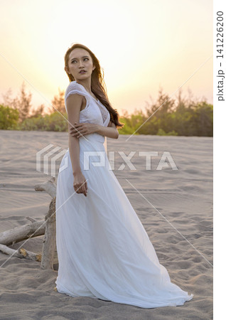 A beautiful young Indonesian southeast asian bride in an elegant white lace wedding gown poses on a sandy beach dunes during a photoshoot, looking away A beautiful young Indonesian southeast asian bride in an elegant white lace wedding gown poses on a sandy beach dunes during a photoshoot, looking away 141226200