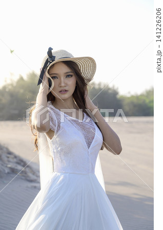 A beautiful young Indonesian southeast asian bride in an elegant white lace wedding gown poses on a sandy beach dunes during a photoshoot, looking away A beautiful young Indonesian southeast asian bride in an elegant white lace wedding gown poses on a sandy beach dunes during a photoshoot, looking away 141226206