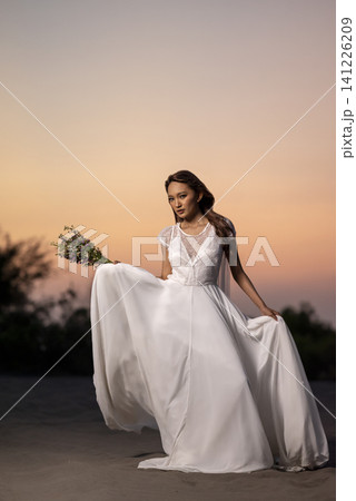 Elegant Indonesian southeast asian bride in a flowing white gown posing on sand dunes during sunset, holding a bouquet 141226209