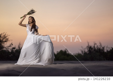Elegant Indonesian southeast asian bride in a flowing white gown posing on sand dunes during sunset, holding a bouquet Elegant Indonesian southeast asian bride in a flowing white gown posing on sand dunes during sunset, holding a bouquet 141226210