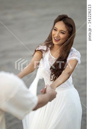 A happy young Indonesian southeast asian woman in a white dress holds hands and dances with her partner on a sandy beach during a romantic pre-wedding or engagement shoot 141226213