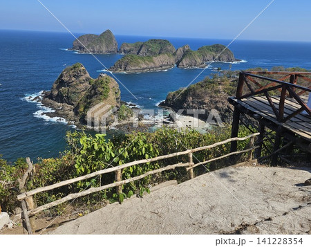 A breathtaking high-angle ocean view from a wooden viewpoint structure, showcasing lush green rocky islets scattered across the vibrant blue sea under the clear sky on a sunny summer day in Indonesia. A breathtaking high-angle ocean view from a wooden viewpoint structure, showcasing lush green rocky islets scattered across the vibrant blue sea under the clear sky on a sunny summer day in Indonesia. 141228354