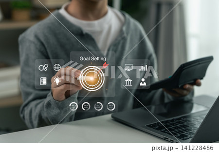 Businessman using virtual interface with digital icons and graphic charts focusing on goal setting concept in modern office while holding calculator and working on laptop computer Businessman using virtual interface with digital icons and graphic charts focusing on goal setting concept in modern office while holding calculator and working on laptop computer 141228486