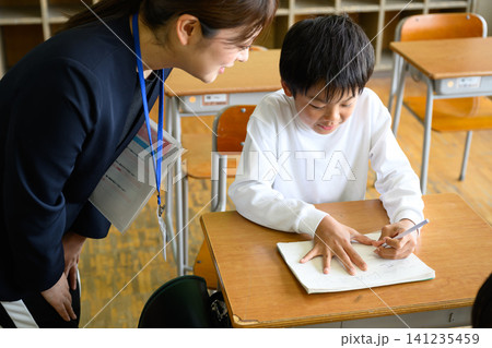 教室の机で勉強する男子小学生を優しく見守り個別指導する女性教師 教室の机で勉強する男子小学生を優しく見守り個別指導する女性教師 141235459