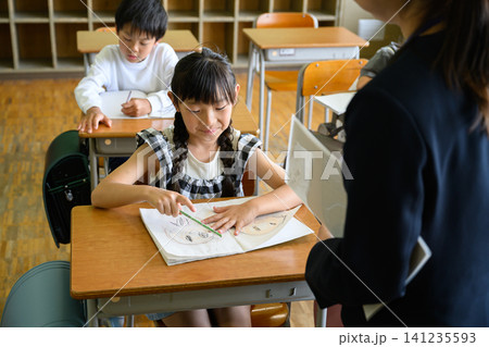 教室で女性教師の個別指導を受けて笑顔でノートを見つめる女子小学生 教室で女性教師の個別指導を受けて笑顔でノートを見つめる女子小学生 141235593