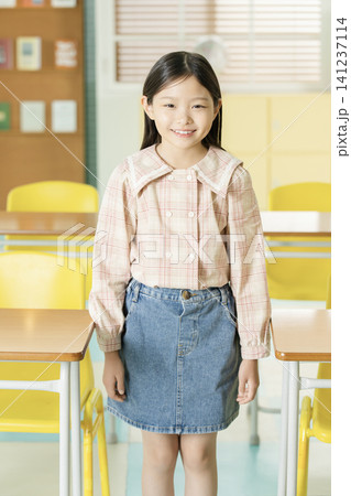 Smiling Asian schoolgirl posing with a backpack between classroom desks Smiling Asian schoolgirl posing with a backpack between classroom desks 141237114