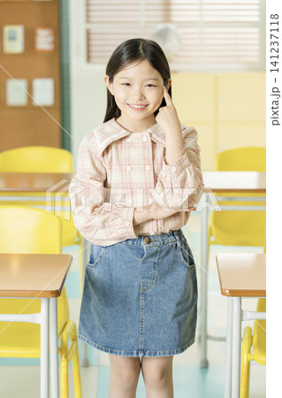 Elementary schoolgirl standing in a classroom with a white backpack 141237118