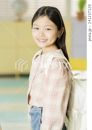 Smiling Asian schoolgirl posing with a backpack between classroom desks Smiling Asian schoolgirl posing with a backpack between classroom desks 141237129