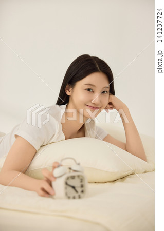 Woman resting on cream bedding beside a vintage alarm clock Woman resting on cream bedding beside a vintage alarm clock 141237724