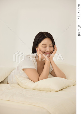 Asian woman lying on a bed and reaching toward an alarm clock 141237726