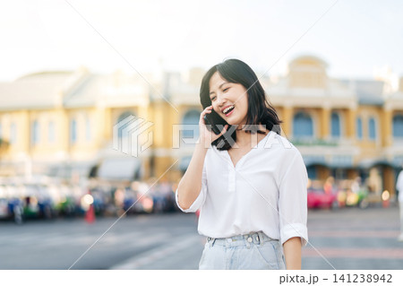 Portrait beautiful young asian woman calling on smart mobile phone standing on outdoor city street. Happy smiling lady talking outside on bright sunny summer day Portrait beautiful young asian woman calling on smart mobile phone standing on outdoor city street. Happy smiling lady talking outside on bright sunny summer day 141238942