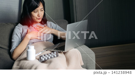 Sick asian woman feeling highly distressed with severe chest pain taking medicine having video call conference medical consultation on laptop while resting comfortably on home sofa blanket Sick asian woman feeling highly distressed with severe chest pain taking medicine having video call conference medical consultation on laptop while resting comfortably on home sofa blanket 141238948