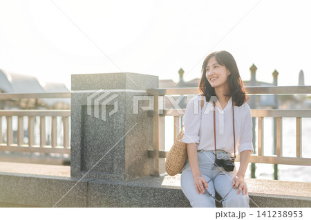 Young beautiful asian woman sitting by river on sunny day with camera enjoying travel vacation trip smiling happily feeling cheerful outdoor leisure lifestyle 141238953