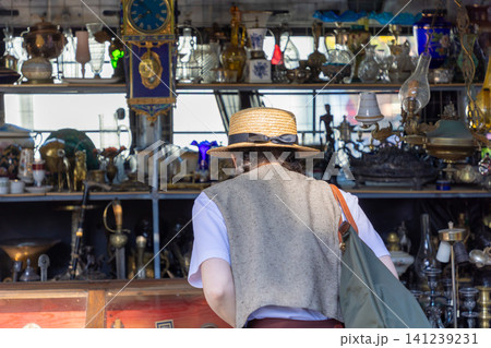 Young woman in a straw hat browsing an antique shop shelf filled with vintage clocks and glassware. Horizontal Young woman in a straw hat browsing an antique shop shelf filled with vintage clocks and glassware. Horizontal 141239231