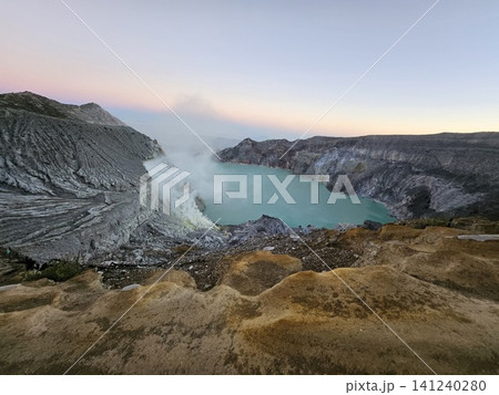 The stunning panoramic sunrise view of the turquoise acid crater lake at Mount Ijen volcano, featuring majestic rocky mountain ridges and thick sulfur smoke rising from the active vents in East Java. 141240280