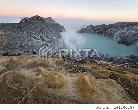 The stunning panoramic sunrise view of the turquoise acid crater lake at Mount Ijen volcano, featuring majestic rocky mountain ridges and thick sulfur smoke rising from the active vents in East Java. 141240287