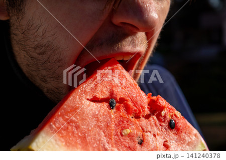 Man eating a fresh slice of watermelon. Close up of male mouth taking a bite of red melon. Summer fruit snack for healthy lifestyle and refreshment. Rural picnic and outdoor food concept. Man eating a fresh slice of watermelon. Close up of male mouth taking a bite of red melon. Summer fruit snack for healthy lifestyle and refreshment. Rural picnic and outdoor food concept. 141240378