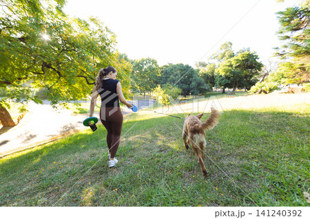 Woman walking a golden retriever dog in a green park, holding a blue frisbee, with trees and sunlight creating a vibrant outdoor scene Woman walking a golden retriever dog in a green park, holding a blue frisbee, with trees and sunlight creating a vibrant outdoor scene 141240392
