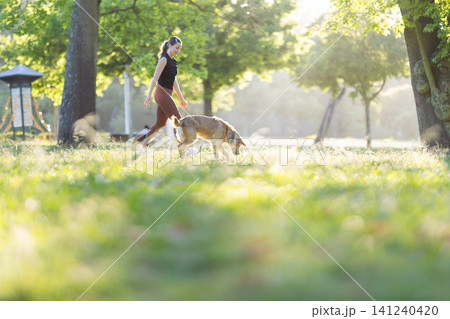 Young woman walking a dog on a sunny day in a park with green grass, trees, and a gazebo visible in the background Young woman walking a dog on a sunny day in a park with green grass, trees, and a gazebo visible in the background 141240420
