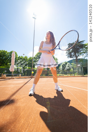 Woman plays tennis on a sunny day at a tennis court surrounded by trees and a clear blue sky Woman plays tennis on a sunny day at a tennis court surrounded by trees and a clear blue sky 141240510