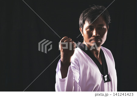 A focused martial artist in a taekwondo uniform raises a clenched fist against a dark background, symbolizing strength, discipline, confidence, and self-defense training A focused martial artist in a taekwondo uniform raises a clenched fist against a dark background, symbolizing strength, discipline, confidence, and self-defense training 141241423