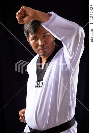 Taekwondo black belt demonstrates high block technique in white dobok against dark studio background, highlighting discipline, defense, and martial arts skill Taekwondo black belt demonstrates high block technique in white dobok against dark studio background, highlighting discipline, defense, and martial arts skill 141241427