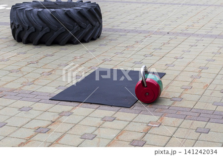 Heavy gym equipment including a red and green kettlebell on a rubber mat and a large tractor tire on a paved outdoor court. Heavy gym equipment including a red and green kettlebell on a rubber mat and a large tractor tire on a paved outdoor court. 141242034