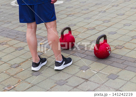 Low section view of an athlete standing on a paved square next to two heavy red 32 kg kettlebells during an outdoor fitness competition. 141242219
