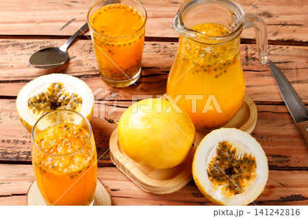 Fresh passion fruit juice in glasses and pitcher with whole and sliced passion fruits arranged on a rustic wooden table with kitchen utensils. Fresh passion fruit juice in glasses and pitcher with whole and sliced passion fruits arranged on a rustic wooden table with kitchen utensils. 141242816
