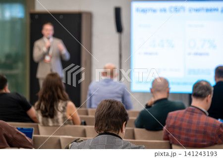 Business audience at conference listening to presentation, blurred speaker and large screen on stage Business audience at conference listening to presentation, blurred speaker and large screen on stage 141243193