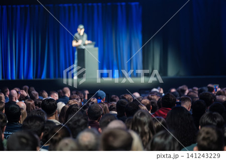 Audience watching presenter on stage during technology conference keynote with blue curtains and spotlight 141243229