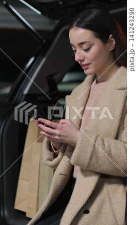 Vertical video Woman using a smartphone while sitting in the trunk of a car in parking lot. Vertical video Woman using a smartphone while sitting in the trunk of a car in parking lot. 141243290