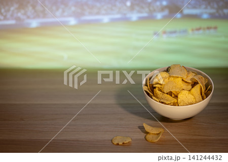 Bowl of potato chips on a table during soccer match broadcast, creating a cozy football night atmosphere with comforting food. Bowl of potato chips on a table during soccer match broadcast, creating a cozy football night atmosphere with comforting food. 141244432